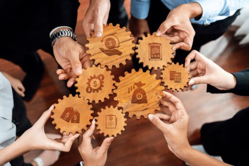 Eine Gruppe von Händen, die ineinander greifende hölzerne Zahnräder mit eingravierten Finanzberatungs- und Geschäftssymbolen halten.