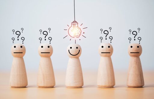 Holzfiguren mit Fragezeichen über dem Kopf, eine Figur lächelt unter einer leuchtenden Glühbirne – Symbol für Erkenntnis und nachhaltige Finanzentscheidungen.