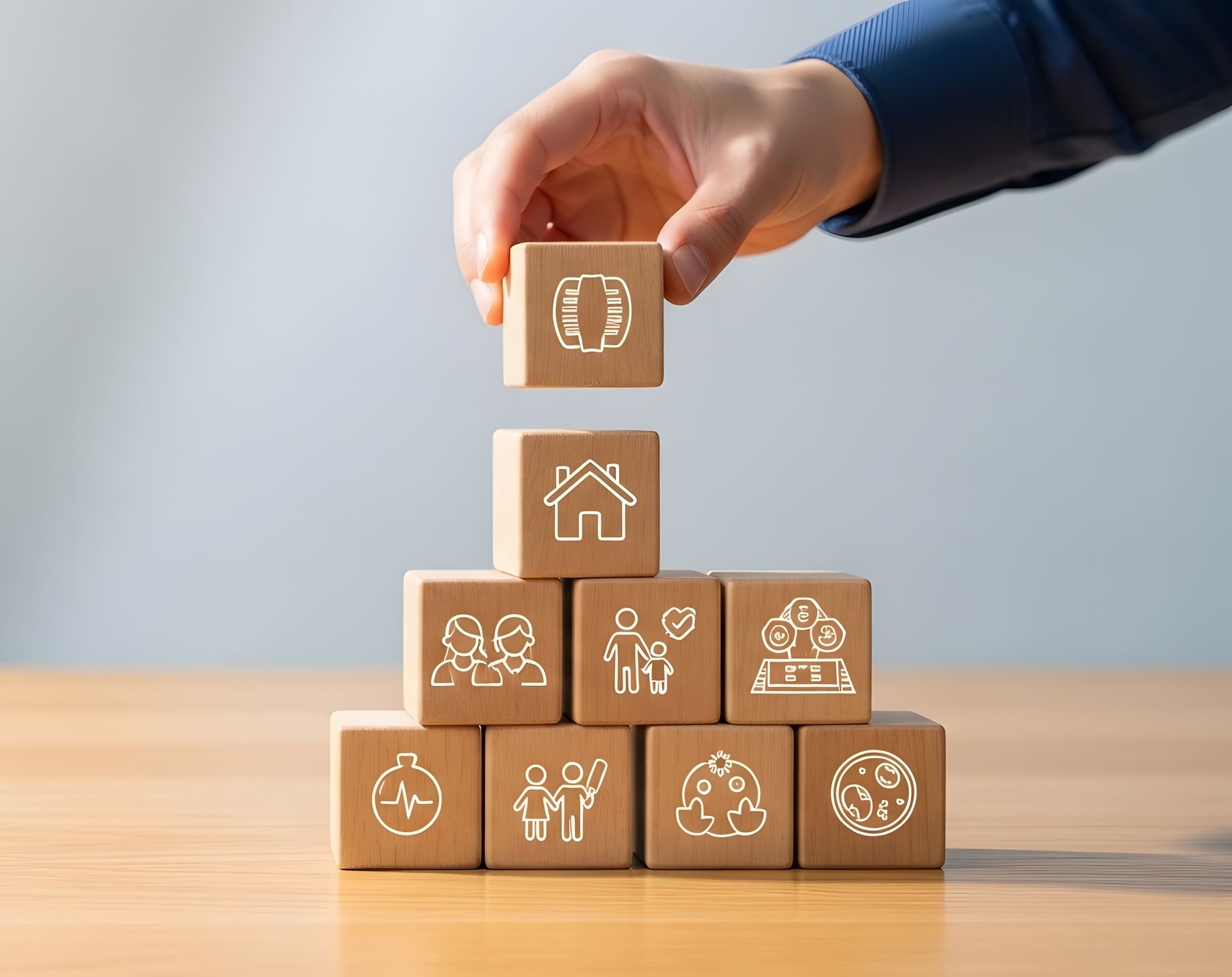 Hand stapelt Holzklötze mit Symbolen für Rente, Haus, Familie, Gesundheit, Vermögen und Schutz.
